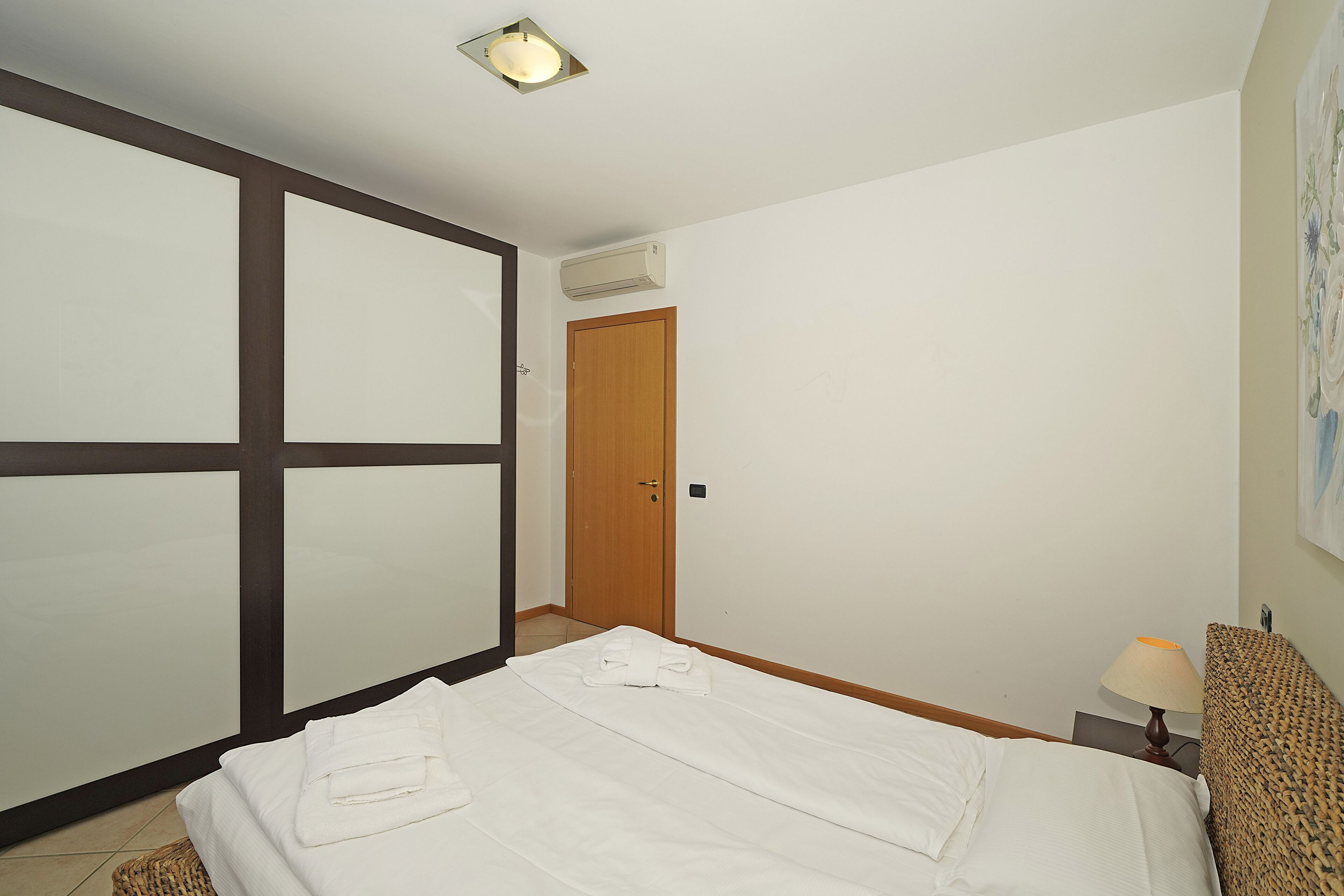 Hotellihuone, jossa on sänky, puinen vaatekaappi, seinälle asennettu ilmastointilaite ja lamppu yöpöydällä.