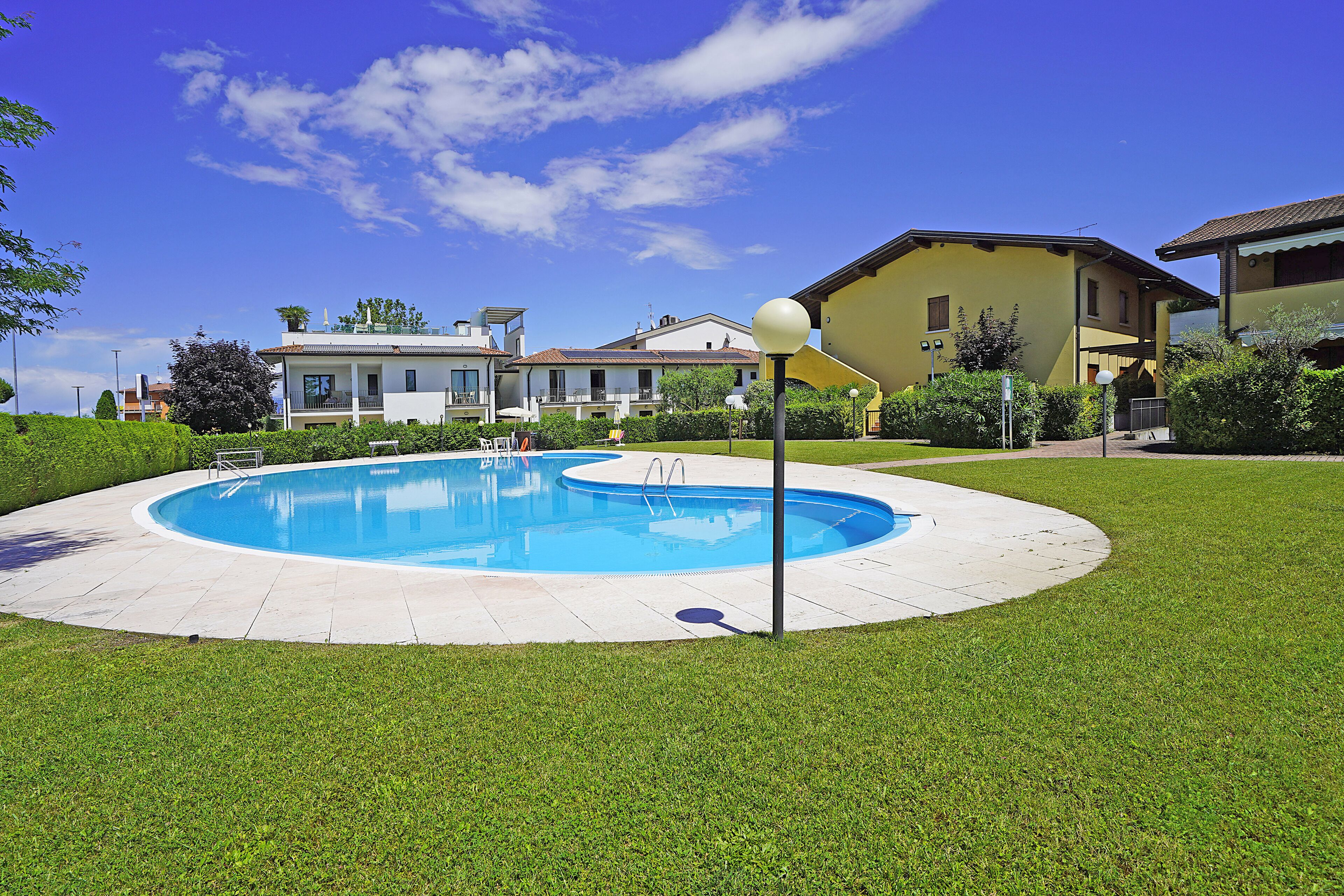 A swimming pool with a clear blue sky, surrounded by well-maintained grass and a few buildings in the background.