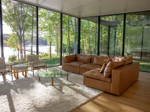 Una sala de estar moderna con un sofá de cuero, una mesa de centro de vidrio y sillas de madera. Amplias ventanas ofrecen vistas a árboles y una masa de agua.
