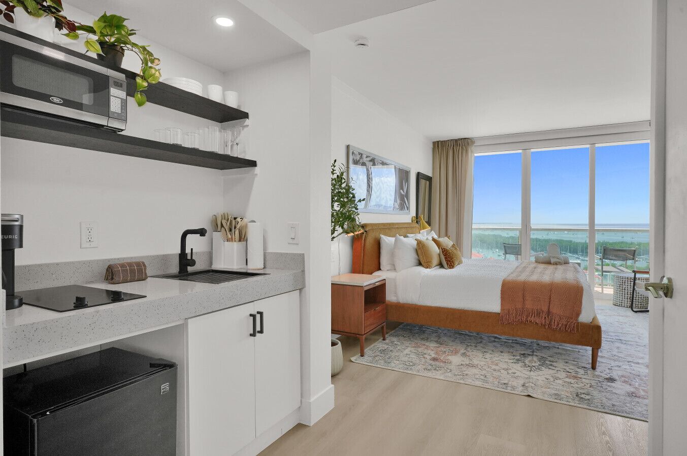 A modern hotel room with a kitchen, a large bed with a view, and a balcony.