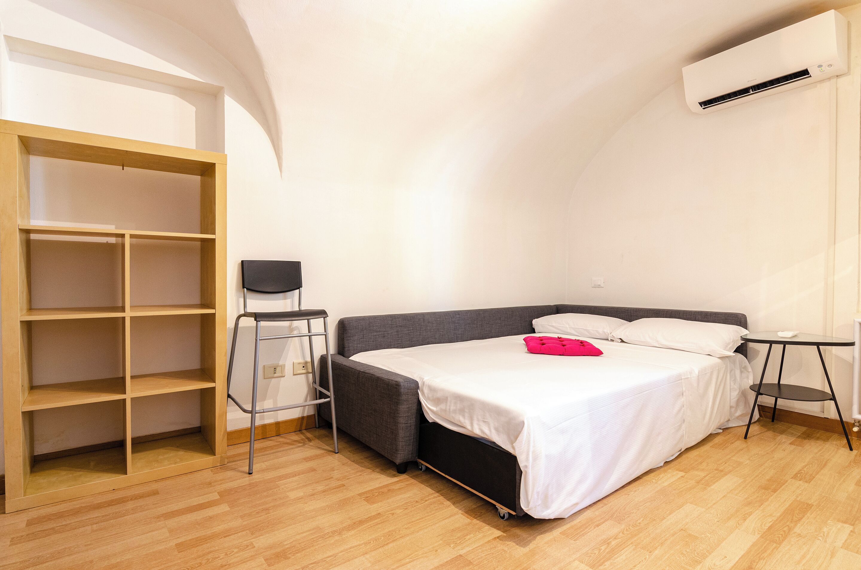 A bedroom with a bed, a small table, a chair, a wardrobe, and an air conditioning unit.