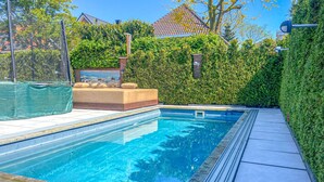 Una piscina con un borde empedrado, un espacio de descanso con cojines y un sendero bordeado de setos.