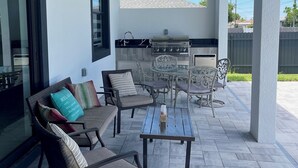 A covered patio with outdoor furniture, a grill, and a view of a house in the background. - Sueños Villa (Cape Coral)