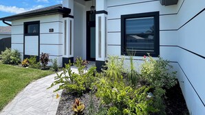 A modern two-story house with a white exterior and black accents, featuring a well-maintained garden with various plants and a paved driveway. - Sueños Villa (Cape Coral)