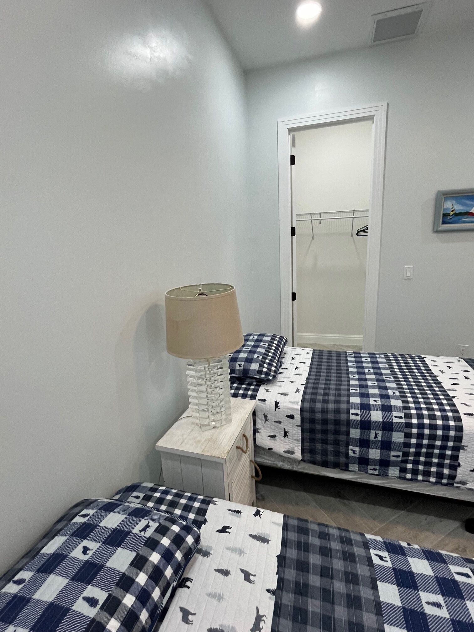 Een slaapkamer met twee bedden, elk met een geruit sprei en bijpassende kussens. Er staat een nachtkastje met een lamp en een klein ladeblok. In de achtergrond is een deur te zien die naar een badkamer leidt.