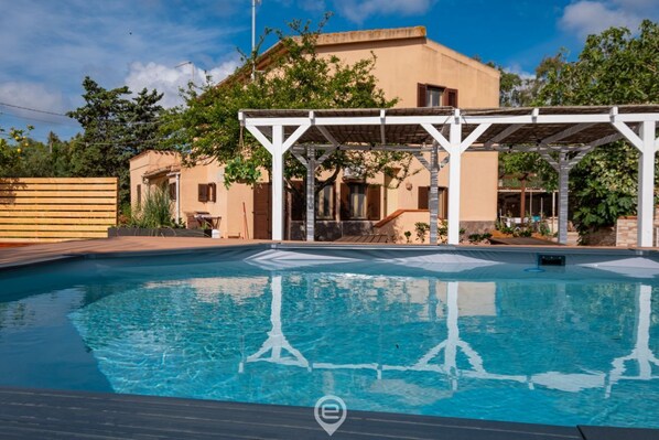 A swimming pool with a pergola structure, a building with a beige facade, and surrounding greenery.