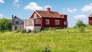 Ein rotes Haus mit weißem Balkon und Schornstein, umgeben von einer grünen Wiese und blauem Himmel.