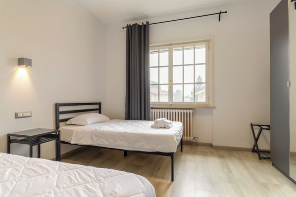 Une chambre avec deux lits, une fenêtre avec des rideaux, un radiateur et un luminaire fixé au mur.
