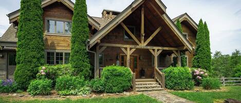A log cabin with a stone pathway leading to the entrance, surrounded by lush greenery and tall trees.