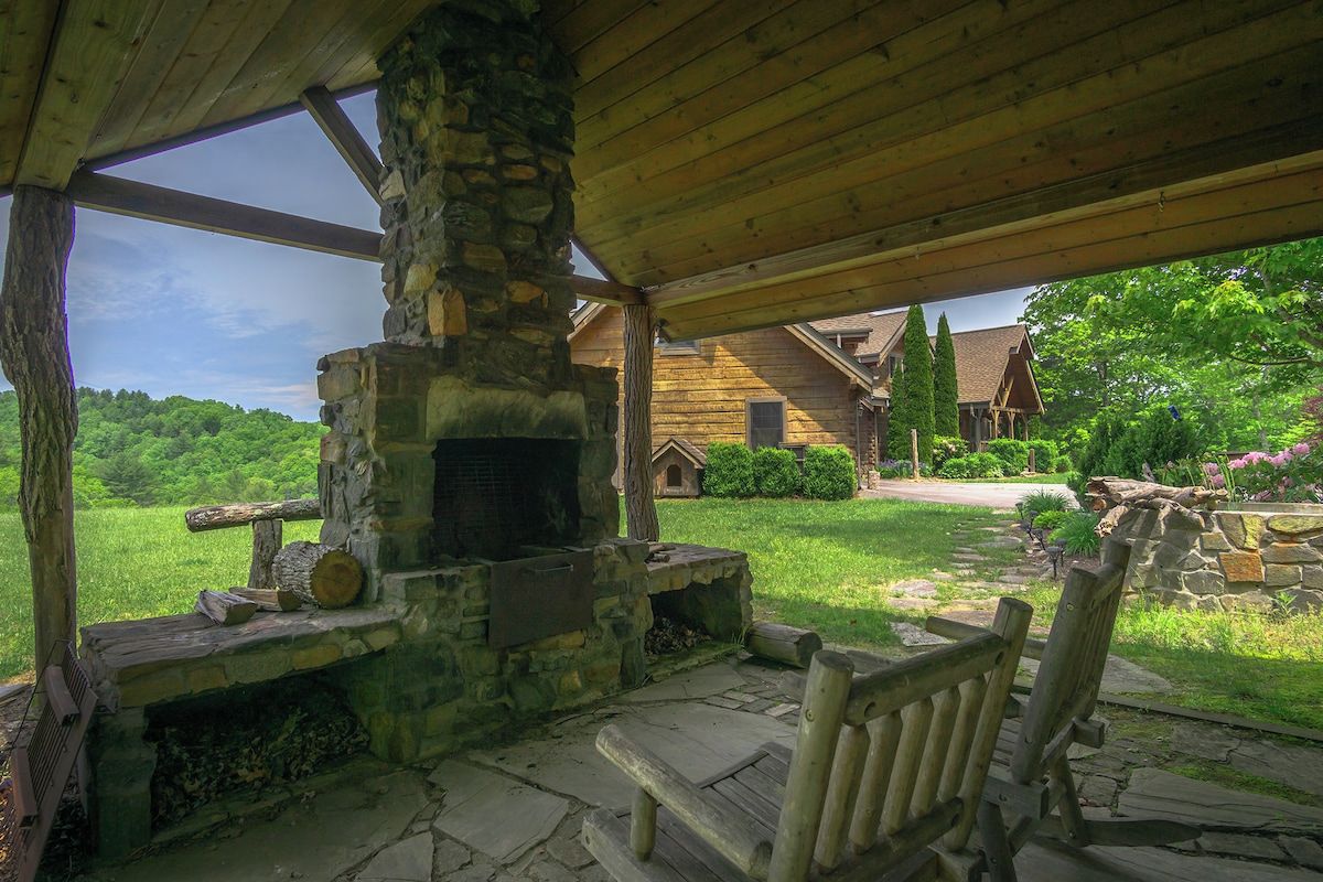 A stone fireplace with a bench in front, set in a covered outdoor area with a view of a wooden house and greenery.