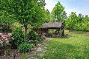 Un bâtiment rustique en pierre, doté d’un porche couvert, entouré d’une végétation luxuriante et bordé d’un sentier en pierre.