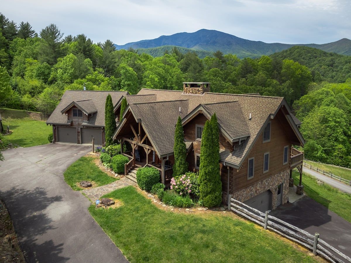 A large, two-story house with a stone foundation and a wooden exterior, surrounded by lush greenery and mountains in the background.