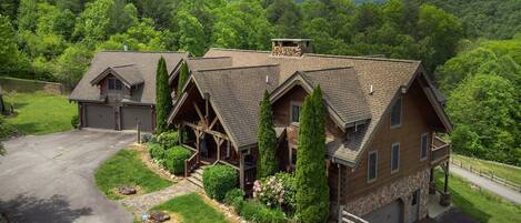 A large, two-story house with a stone foundation and a wooden exterior, surrounded by lush greenery and mountains in the background.