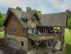 Une cabane en rondins à deux étages, avec des fondations en pierre, une terrasse couverte dotée de meubles d’extérieur et un garage.