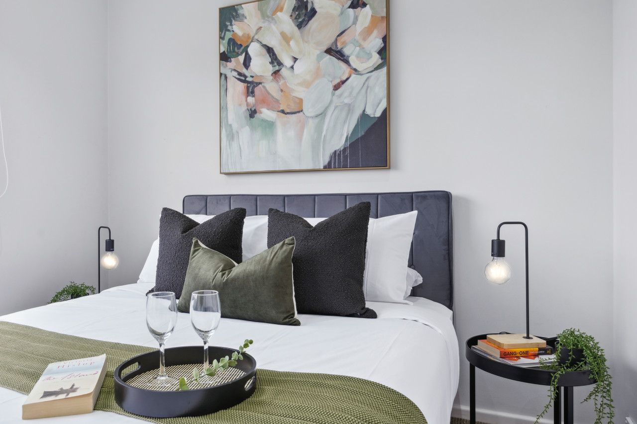 A neatly arranged bedroom with a bed, pillows, bedside tables, a wall art piece, and a tray with glasses and a book.