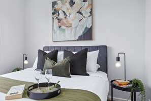 A neatly arranged bedroom with a bed, pillows, bedside tables, a wall art piece, and a tray with glasses and a book. - The Fulton Residence (Saint Kilda East)