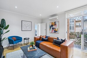 A modern living room with a leather sofa, a blue armchair, and a coffee table. There is a large window providing a view of the outside. - The Fulton Residence (Saint Kilda East)