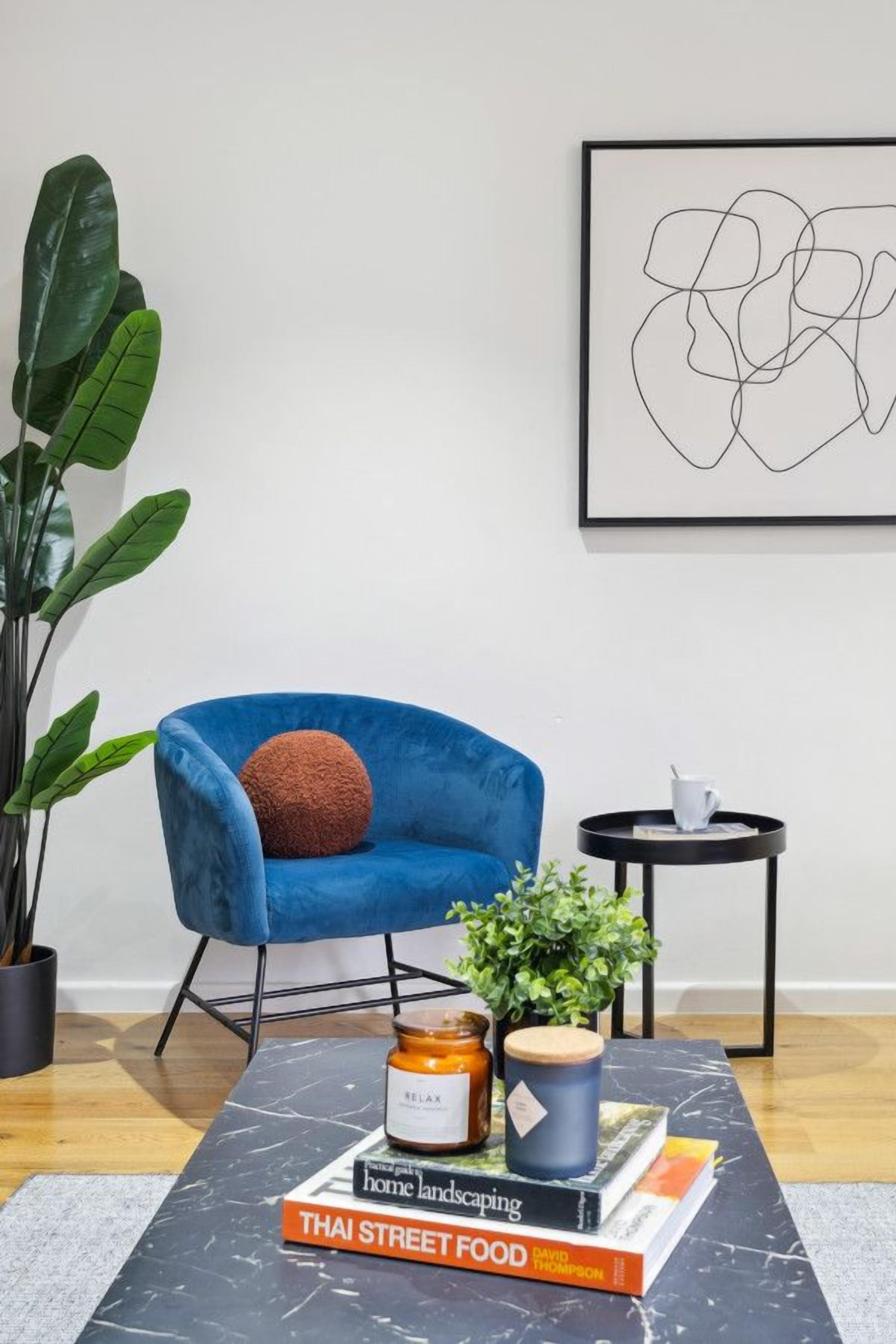 A modern living room with a blue armchair, a coffee table with books, a small side table, and a large green plant.