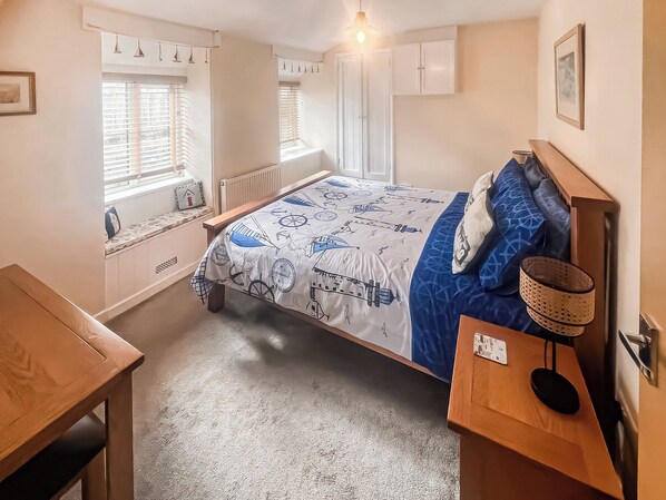 Une chambre à coucher comprenant un lit, un bureau, une chaise, une fenêtre avec des stores, un radiateur et une étagère fixée au mur.