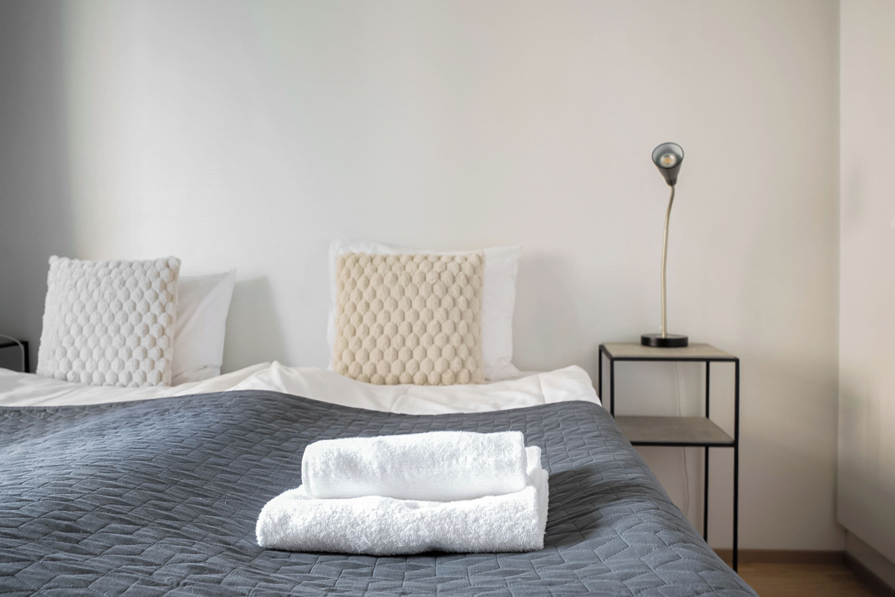 Ein ordentlich gemachtes Bett mit einer grauen Tagesdecke, zwei weißen Kissen mit einer strukturierten Oberfläche und einem gefalteten weißen Handtuch.