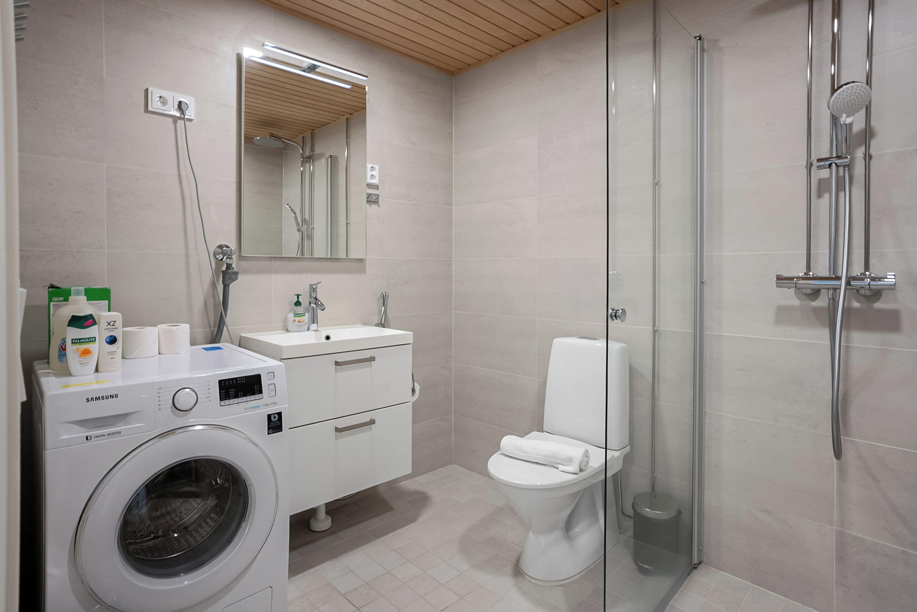 A modern bathroom with a washing machine, a sink, a toilet, and a glass-enclosed shower area.