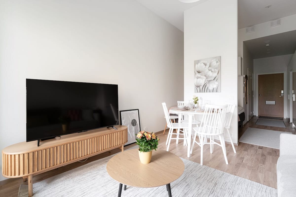 Ein modernes Wohnzimmer mit Flachbildfernseher, einer Holzkonsole, einem Essbereich mit weißen Stühlen und einem runden Tisch mit Topfpflanze.