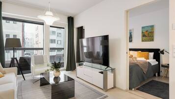 A modern hotel room with a large TV, a sofa, a glass coffee table, and a bed with a black headboard.