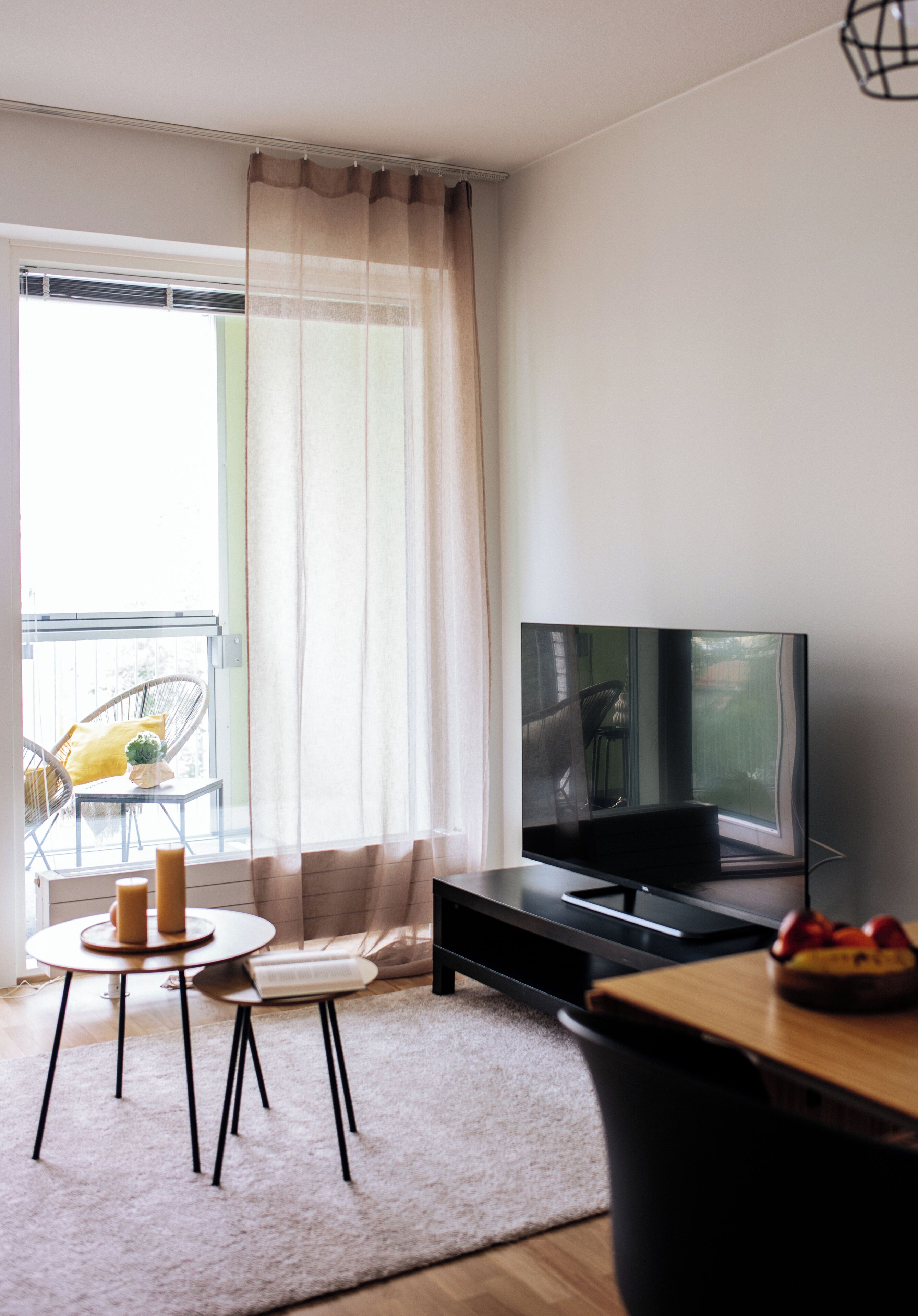 Ett modernt vardagsrum med en platt-TV, ett litet runt bord med ljus och en stol vid ett fönster med tunna, genomskinliga gardiner.