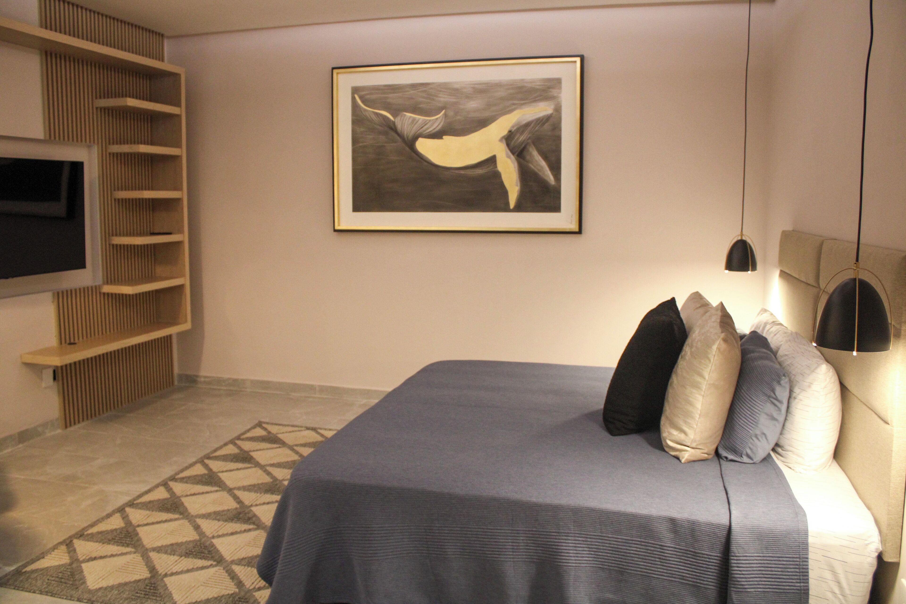A modern bedroom with a bed, pillows, a wall-mounted TV, a framed artwork, and a patterned rug.