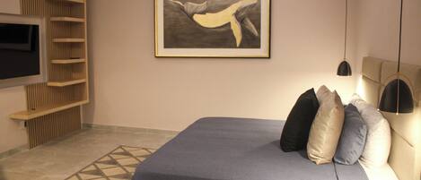 A modern bedroom with a bed, pillows, a wall-mounted TV, a framed artwork, and a patterned rug.