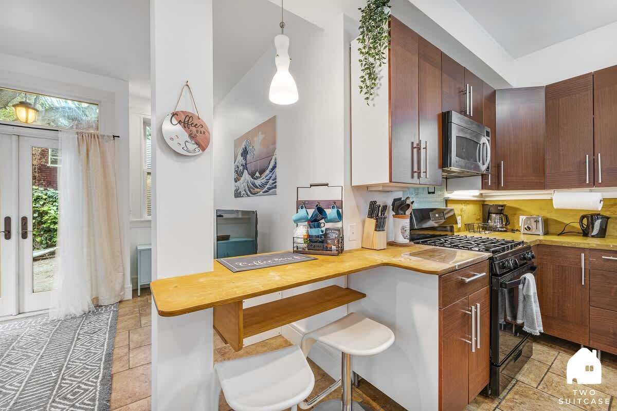 A compact kitchen with wooden cabinets, a microwave, a stove, and a sink. There are two stools and a hanging light fixture.