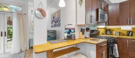 A compact kitchen with wooden cabinets, a microwave, a stove, and a sink. There are two stools and a hanging light fixture.
