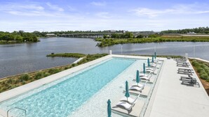 A rectangular pool with clear water, surrounded by lounge chairs and umbrellas, with a view of a river and bridge in the background. - Brand New 2 Bedroom Condo-Luxurious Waterfront Pool-Sleeps 4 (Gulfport)