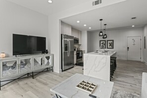 A modern kitchen with a built-in refrigerator, microwave, and oven. There is a dining area with a marble table and chairs. A flat-screen TV is mounted on the wall.