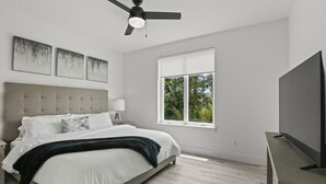 A modern bedroom with a large bed, a ceiling fan, a TV on a stand, and a window with a view of trees. - Brand New 2 Bedroom Condo-Luxurious Waterfront Pool-Sleeps 4 (Gulfport)