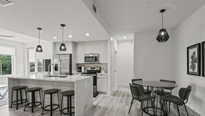 A modern kitchen with a central island, bar stools, and a dining area with a round table and chairs. - Brand New 2 Bedroom Condo-Luxurious Waterfront Pool-Sleeps 4 (Gulfport)
