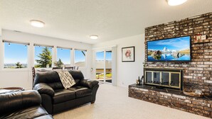 A living room with a fireplace, a black leather sofa, and a television mounted on a brick wall. - Red Hills Vacation | 3BR Hot Tub, Sauna & Views (Dundee)
