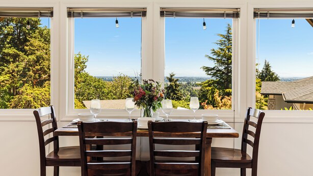 A dining table with four chairs, set for a meal, positioned in front of a window with a view of trees and a distant landscape. - Red Hills Vacation | 3BR Hot Tub, Sauna & Views (Dundee)