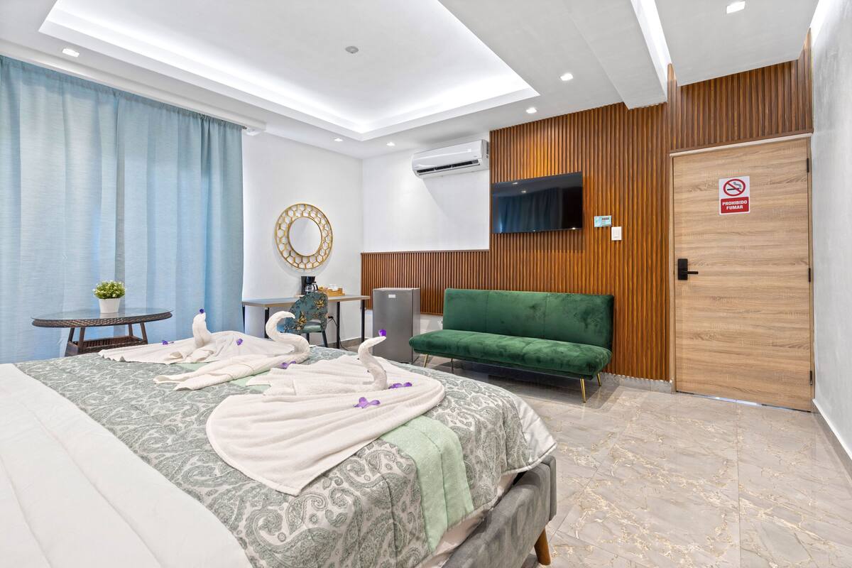 Een moderne hotelkamer met een groot bed, een groene bank, een klein tafeltje met een plant en een aan de muur bevestigde televisie.