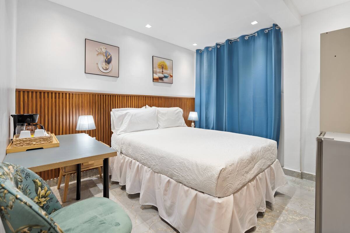 Een hotelkamer met een houten hoofdeinde, een bed met witte lakens, een bureau met een lamp, een stoel en een blauw gordijn.