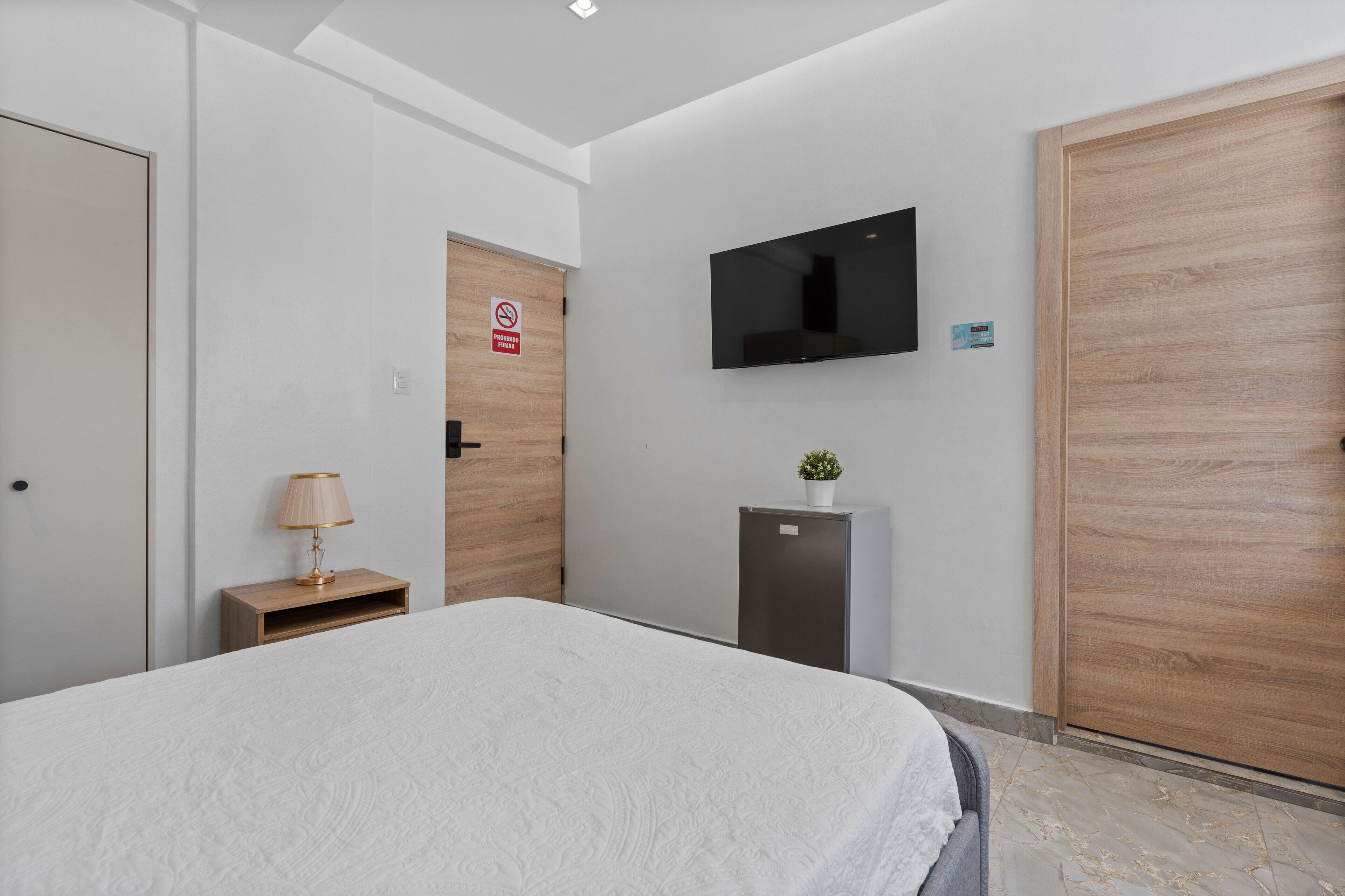 A modern hotel room with a large bed, a television mounted on the wall, a small desk with a lamp, and a door with a no-smoking sign.