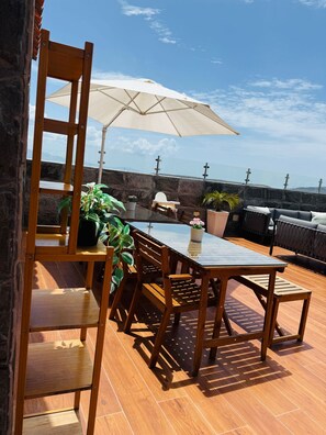 Una terraza de madera con una mesa, sillas, una sombrilla grande y macetas con plantas.