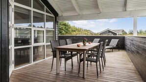 Eine überdachte Terrasse mit Holztable und -stühlen, großen Fenstern und Blick auf die Umgebung.