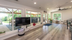 A spacious living room with a large flat-screen TV, wooden flooring, and large windows offering a view of the outdoors. - Gorgeous Modern Lakeside Getaway in Gun Barrel City (Gun Barrel City)