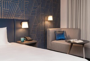 A hotel room with a bed, a nightstand with a phone and a mug, a seating area with a chair and a table, and a patterned wall. - Hyatt Place Leeds (Leeds)