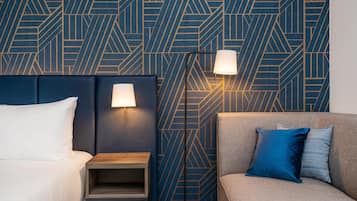 A hotel room with a geometric patterned wall, a bed with white bedding, a bedside table, a lamp, and a sofa with blue and grey cushions.