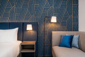 A hotel room with a geometric patterned wall, a bed with white bedding, a bedside table, a lamp, and a sofa with blue and grey cushions. - Hyatt Place Leeds (Leeds)