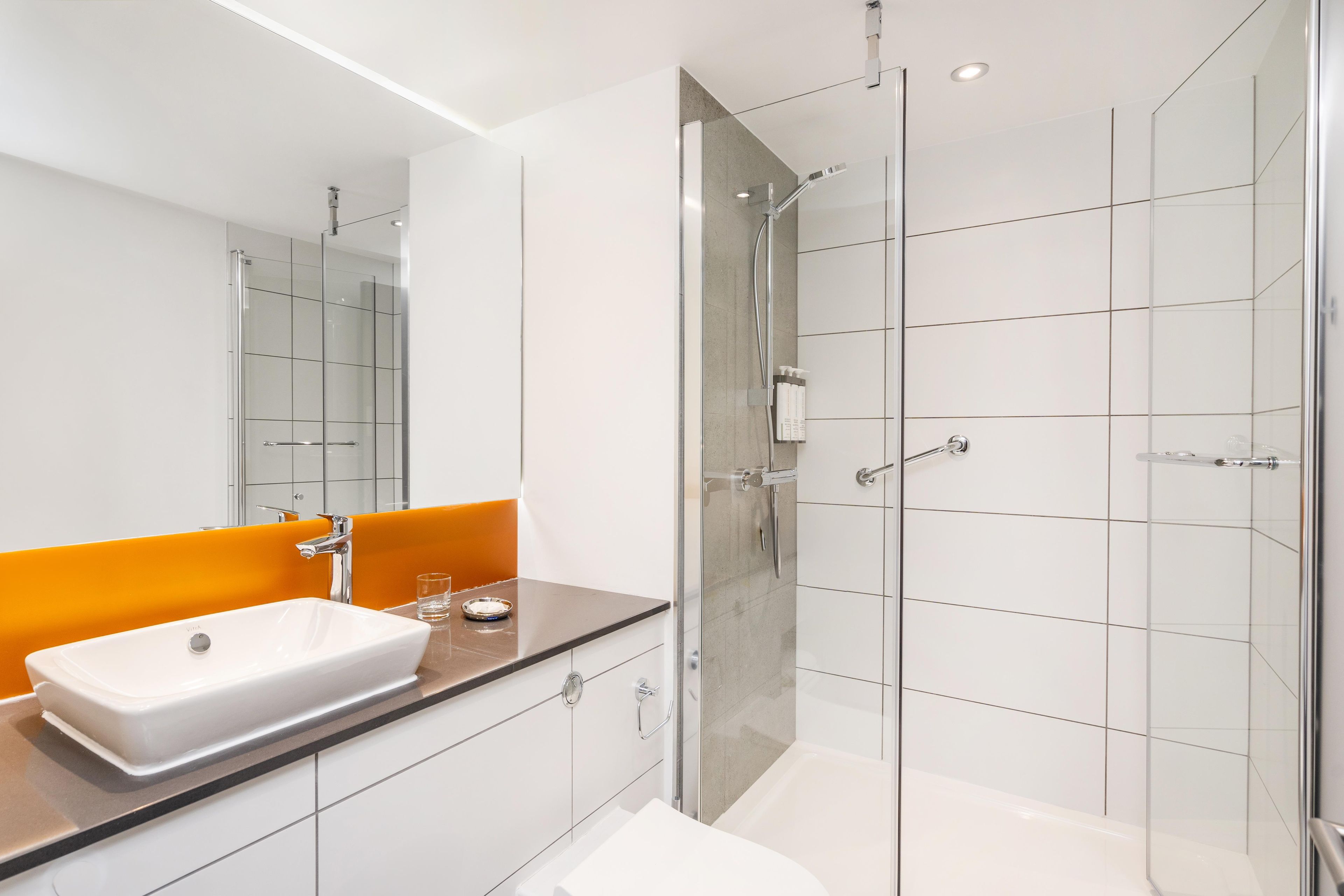 A modern bathroom with a glass shower enclosure, a white sink, and an orange accent wall.