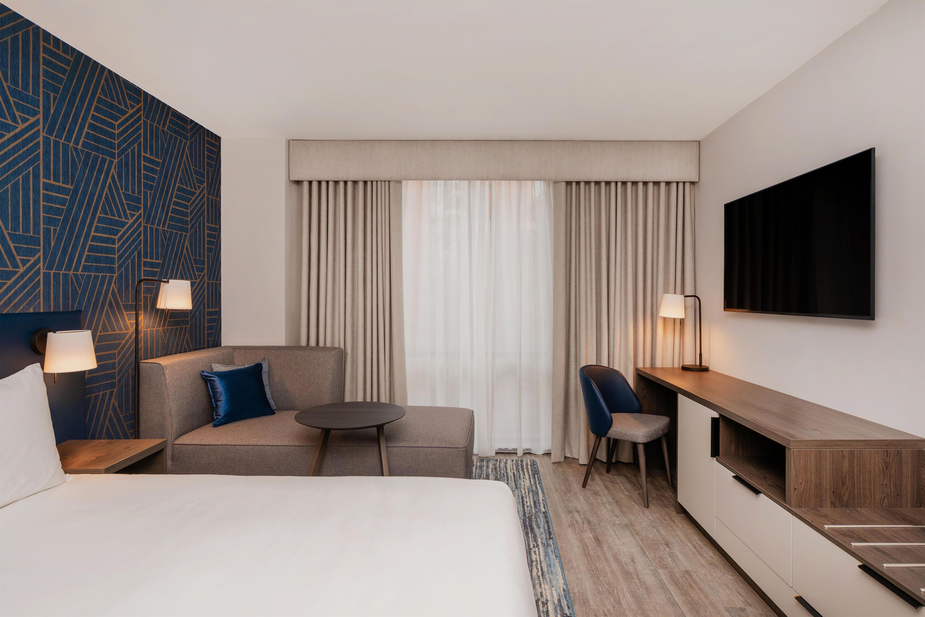 Een hotelkamer met een bed, een bank, een klein tafeltje, een stoel, een bureau, een televisie en gordijnen.