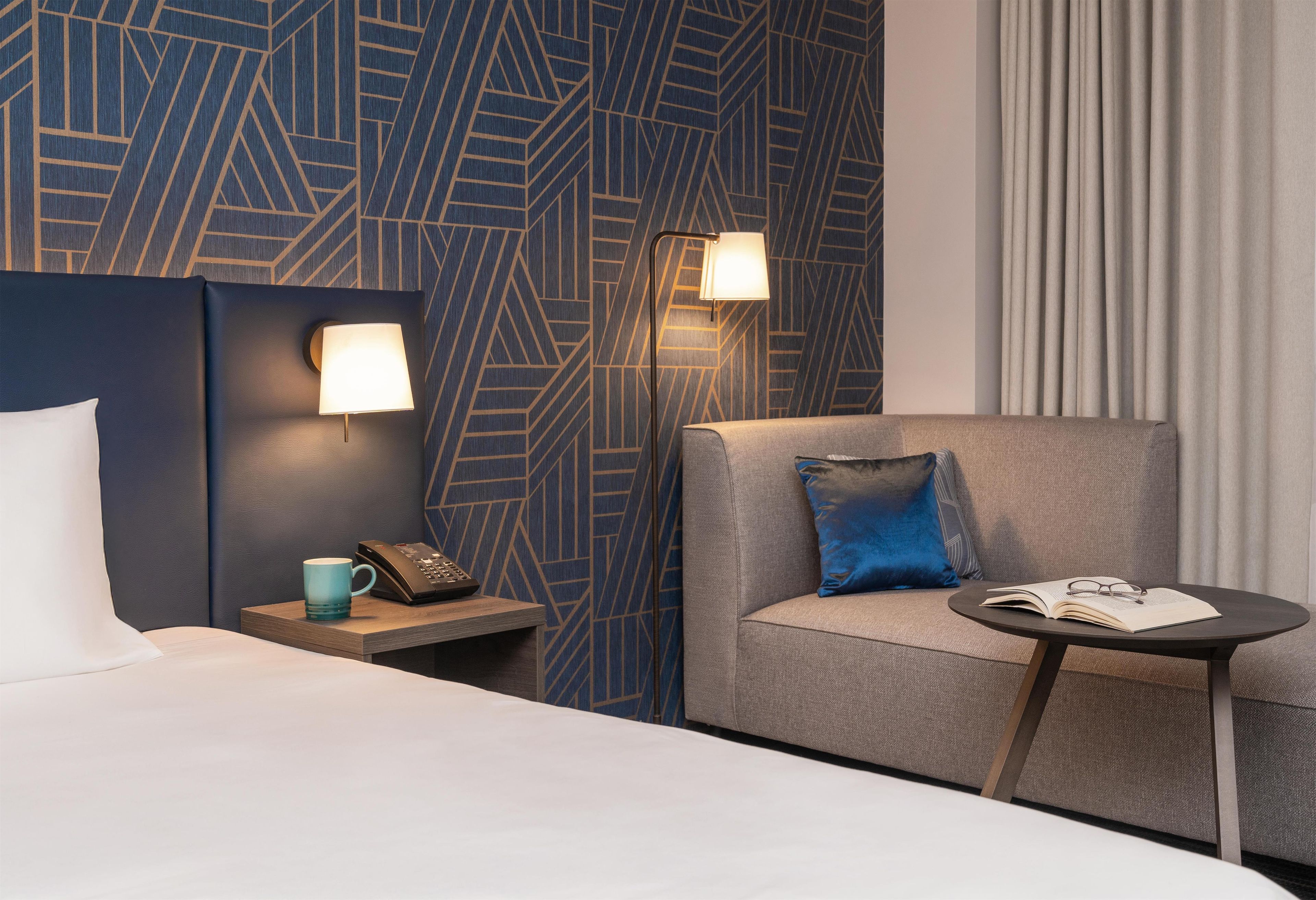 Una habitación de hotel con una cama, una mesita de noche con un teléfono y una taza, un espacio de estar con una silla y una mesa, y una pared con un diseño estampado.